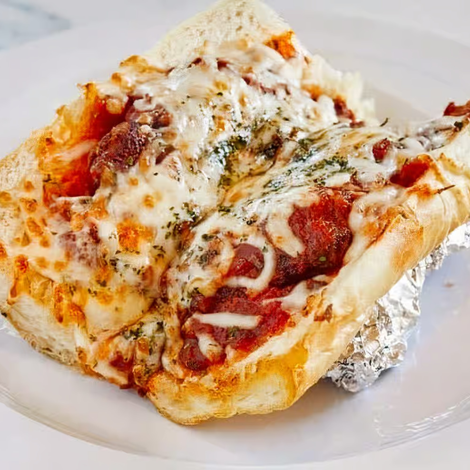 A cheesy meatball sub sandwich with marinara sauce and herbs on a white plate.